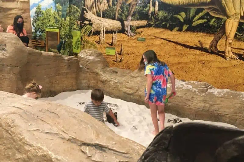kids in a sandbox with t-rex legs in the background