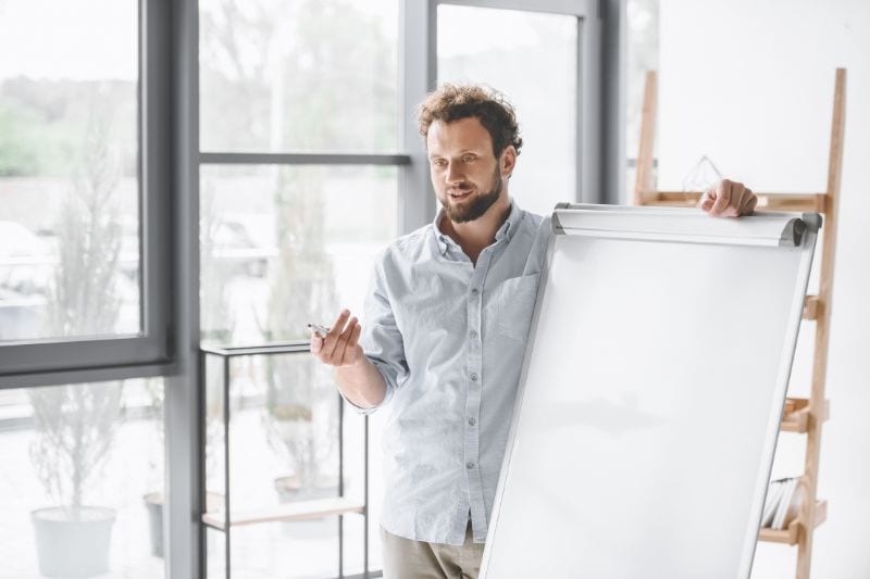 man in button down shirt standing at a white board speaking