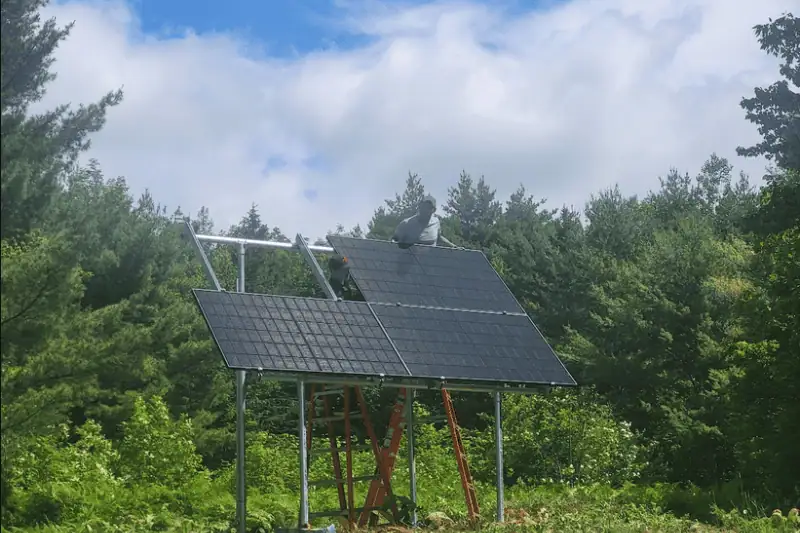 The image shows two people installing solar panels on a metal frame structure in a natural, outdoor setting. The panels are tilted and mounted on a raised platform supported by poles, with ladders leaning against the frame for access. One person is standing on top of the panel array, while the other is partially visible behind it, both appearing to work on the installation.

The background features dense green trees under a partly cloudy blue sky, suggesting a rural or forested location. The scene conveys a sense of sustainable energy development in harmony with nature.