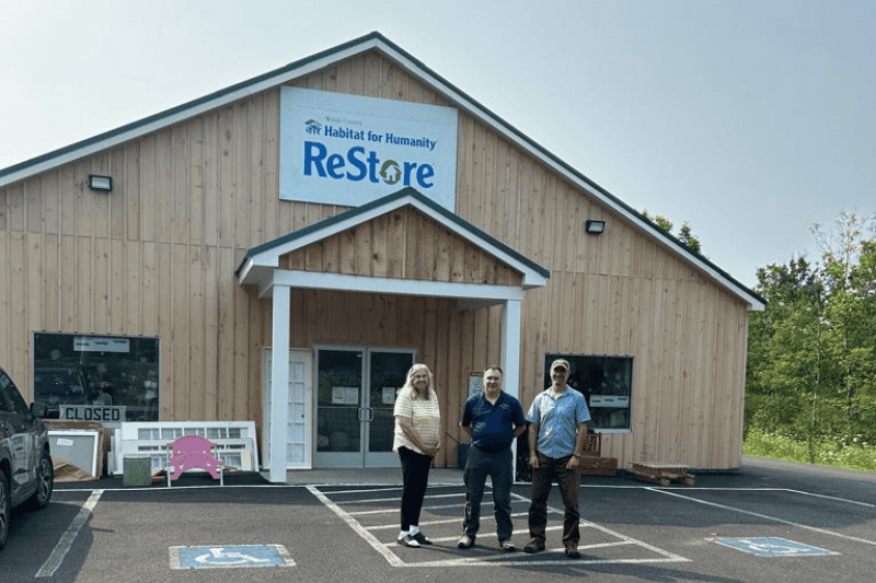 a photo taken in front of Habitat for Humanity Restore in Waldo County. the building is made from white wooden planks with a ReStore sign in blue letter and a white background behind it. In this picture 3 people stand in front of the building: a lady in yellow t-shirt with long white hair, Richard Marino, Everybody Solar board member in a dark blue shirt and black pants and Vice President of Restore Todd Bluhm in a light grey shirt, black pants and grey cap on his head. 
