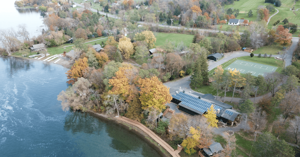 A picture of a building from above, like from a drone, with solar panels sitting on the roof. The building is located on the lake and is surrounded by autumn trees. 