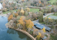 A picture of a building from above, like from a drone, with solar panels sitting on the roof. The building is located on the lake and is surrounded by autumn trees. 